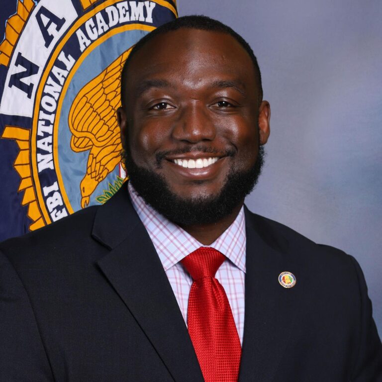 A portrait of Jacksonville police Chief Marcus Wood smiling in front of a flag with the logo of the Alabama Association of Chiefs of Police.