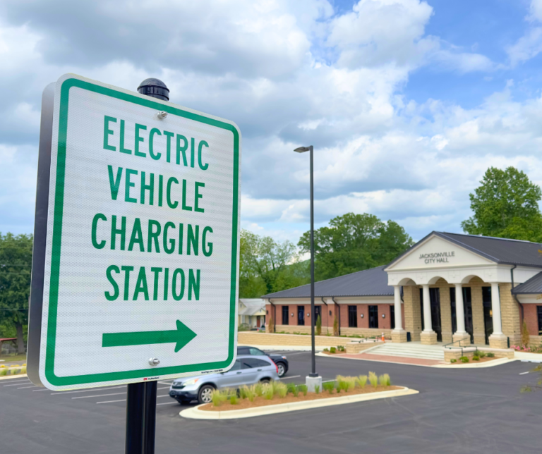 Street sign that reads "electric vehicle charging station" with an arrow pointing into the Jacksonville City Hall parking lot.